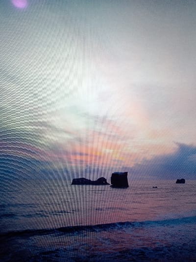 This image depicts a coastal seascape at either dawn or dusk, located at the coast of Olambe, Nigeria. The upper portion displays a sky gradient, transitioning from light gray at the top to soft blues, pinks, and oranges at the horizon. A distinct purple light anomaly is present in the upper left sky. The lower portion features dark ocean water, reflecting the sky's colors, with subtle wave patterns visible in the immediate foreground. Several dark, prominent rock formations, likely sea stacks or islets, are observed within the water: an elongated, multi-humped formation left of center, a taller, block-like formation adjacent to it on the right, and two smaller, less defined formations further in the distance towards the right. No people or specific human activities are visible within the frame. The entire image is overlaid with a uniform, distinct grid pattern, characteristic of a photograph taken of a digital display screen.