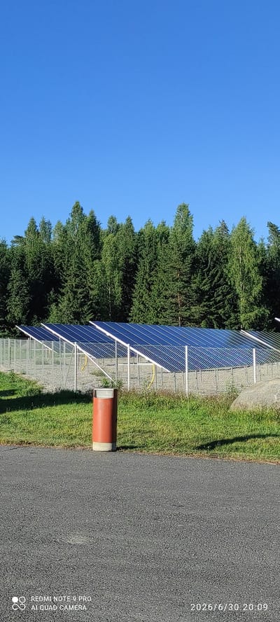 This image shows a solar panel array situated in an outdoor setting, likely a rural area, given the presence of lush green trees and a clear blue sky. The solar panels are mounted on a white metal frame and are angled to face the sun. They appear to be a dark blue or black color.
Adjacent to the solar panels is a well-maintained grassy area, suggesting that the area is regularly cared for. A large, rough-textured grey rock is partially visible in the grassy area, adding a natural element to the scene. A dark grey asphalt surface runs along the edge of the grass, indicating a pathway or road.
In the foreground, a cylindrical object with a red exterior and a grey concrete base stands upright. This object might be part of the solar panel installation, perhaps a protective casing or a marker. A chain-link fence runs along the front of the solar panels, potentially for security or to delineate the area.
The lighting suggests it's daytime, with bright sunlight casting shadows. The clear blue sky indicates fair weather. The overall scene conveys a sense of a modern, sustainable energy installation integrated into a natural environment.
Visible text includes: "2026/6/30 20:09" and "O REDMI NOTE 9 PRO AI QUAD CAMERA". The former is likely a timestamp, and the latter is a camera watermark.
The location context is Joutsa, Finland.