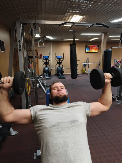 A male subject with a dark beard and wearing a light grey, sweat-stained t-shirt is performing a dumbbell press while lying on a blue and grey weight bench. He holds a black dumbbell in each hand, with arms extended upwards. The scene is set in a gym in the city of Im. Gastello, Russia. The facility features dark red/brown rubberized flooring and light brown/tan panelled walls. The ceiling is a grid system with integrated fluorescent lighting fixtures. In the background, various gym equipment is present, including a silver "FOREMAN" brand cable machine, two black elliptical trainers with illuminated blue screens, a black punching bag suspended from the ceiling, a barbell rack with a barbell and yellow weight plates, and a wall-mounted television displaying a vibrant, colorful image or video.