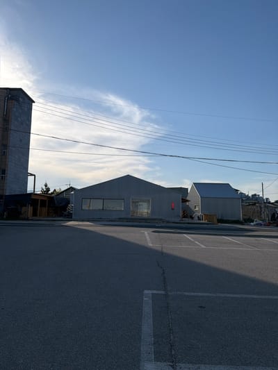 The image captures an urban scene in Toseong-myeon, South Korea. A modern, light gray building with a gabled roof and large windows stands along a paved road or parking area marked with white parking lines. Multiple utility wires stretch across the blue sky above the buildings. Adjacent to the gray building is what appears to be a small, makeshift structure, possibly a shed or temporary shelter. Further in the background, other buildings, including a blocky, light-colored building, rise up on the opposite side of the street. The asphalt road surface is in the foreground, and the overall light indicates it is daytime.