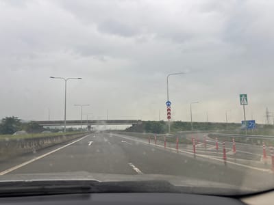 The image depicts a view from a vehicle driving on a wet highway in the municipality of Nigotits Temi, Georgia. The sky is overcast with dense clouds, suggesting a gloomy or rainy day. The highway has multiple lanes, marked with white lines and directional arrows. Several streetlights line the sides of the road. In the distance, an overpass spans the highway, and on the right side of the road, there are traffic signs, including one with green and white markings indicating an exit or route, and another blue sign with directional arrows. Orange traffic cones with white reflective stripes are placed along the right lane, potentially indicating a closed lane or construction. The roadside is lined with a concrete barrier and grassy slopes with some vegetation. Several vehicles are visible on the highway, and some cars are parked on the side of the road beyond the concrete barrier. The overall environment appears to be a rural or semi-rural highway setting.