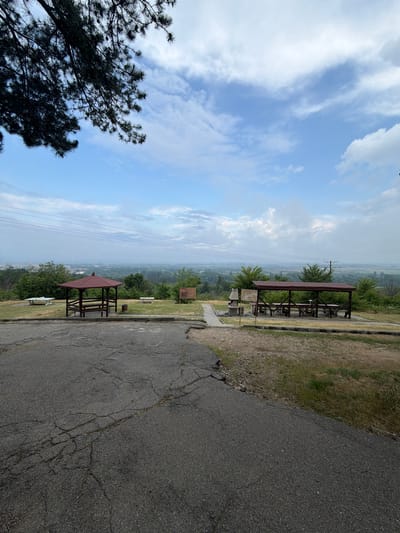 The image captures a peaceful outdoor scenic viewpoint, likely a rest stop or park, under a partly cloudy daytime sky. The scene is set in Kran, Bulgaria.
In the foreground, a dark asphalt road with visible cracks occupies the right side of the frame, curving gently away. Adjacent to the road on the left, there's a patch of dry, sparse grass and dirt that transitions into a greener area further back. A prominent, dark-silhouetted pine-like tree with dense needles and visible cones frames the bottom-left corner of the image, contrasting sharply with the bright sky.
In the midground, two identical wooden picnic shelters are positioned on a grassy area overlooking a distant landscape. Each shelter features a reddish-brown, sloped roof and includes picnic tables and benches underneath. One shelter is closer to the road, and another is further back and slightly to the left. A small wooden sign is visible near the closest shelter, but its text is illegible. Utility poles and power lines stretch across the mid-distance.
The background presents a wide, hazy panoramic view of a valley, densely covered with trees and suggesting a distant town or settlement beneath the vast expanse of a bright blue sky dotted with scattered white clouds. The overall atmosphere is tranquil. No individuals are present in the scene. The time of day appears to be late morning or early afternoon, with ample natural light.