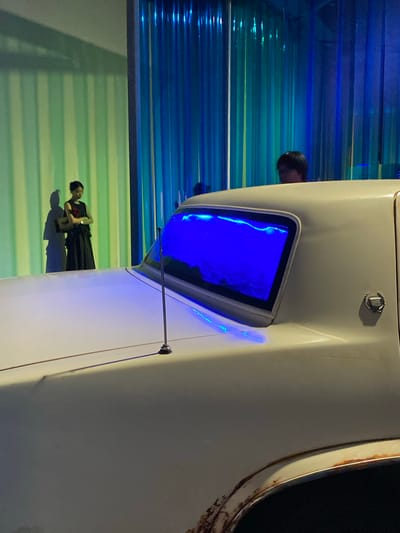 In Jinniu District, China, a white car with rusted accents is positioned indoors. The car's rear window glows with a vibrant blue light, creating an ethereal atmosphere. Behind the car, a woman stands with her arms crossed, dressed in a black sleeveless dress with a red floral accent on the bodice and a long black skirt. Her shadow is cast on a striped green and light green wall. To the right of the woman, a person with dark hair is visible behind the car, partially obscured by its roof. The background is characterized by vertical blue and teal curtains, illuminated by colored lights, suggesting an artistic exhibition or performance space.