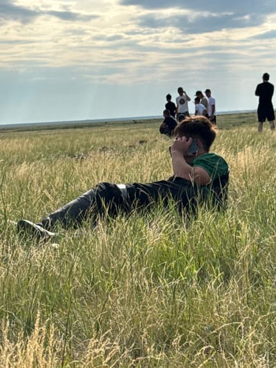The image depicts a man lying in tall grass, holding a cell phone to his ear. He is wearing a green shirt and dark pants. In the background, a group of people are standing and looking in the distance. The environment appears to be a vast, grassy plain under a cloudy sky. The location is identified as the city of Өндіріс, Kazakhstan. The scene is open and rural, with no visible urban structures. The lighting suggests it is daytime, likely late afternoon or early evening due to the soft, diffused light.