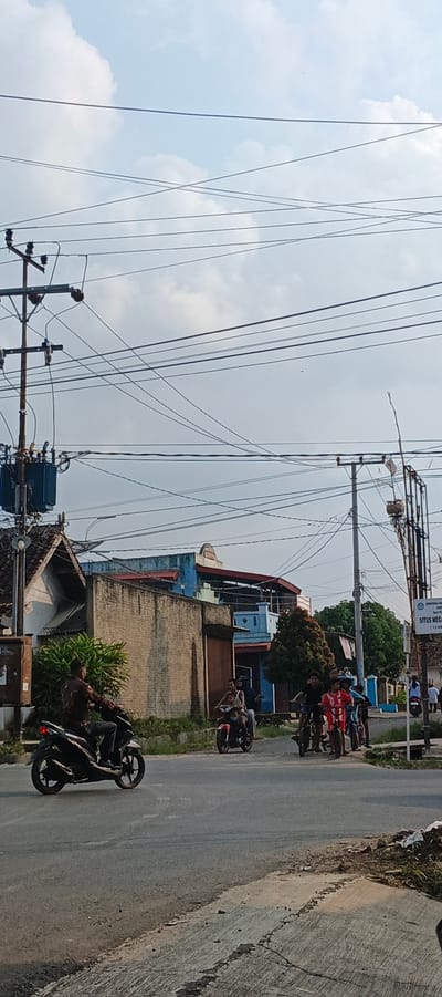 The image depicts a street scene in Sukarame, Indonesia, characterized by a network of power lines crisscrossing a cloudy sky. In the foreground, a dark-colored motorcycle, ridden by a person wearing a patterned long-sleeved shirt and gray pants, travels on an asphalt road. To the right, a group of individuals on bicycles and a red motorcycle are also present on the road. Behind the road, a low-rise building with a textured brick wall and a pitched roof is visible, adjacent to a structure with blue and red facades. A blue transformer box is mounted on a utility pole on the left side of the image. A signpost with text, partially obscured, stands on the right. The environment appears to be a suburban or semi-urban area.