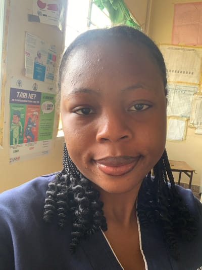 The image is a close-up selfie of a young Black woman with braided hair, wearing a dark blue uniform with a white trim. She is looking directly at the camera with a slight smile. The background suggests an indoor setting that appears to be a clinic or office. Posters are visible on the wall behind her, including one with the text "TARI NE?" and cartoon illustrations, which is likely health-related educational material. Another poster shows a chart with rows and columns, possibly a data or record-keeping chart, and the WHO logo is partially visible. The lighting in the room comes from a window on the left, which is partially covered by green fabric. A wooden table and chair are visible in the background on the right. The overall impression is that of a casual, informal photograph taken within a healthcare or public service facility, likely in Reak, Nigeria, given the context.