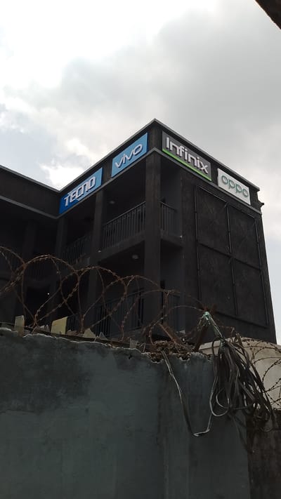 A two-story dark-colored building stands behind a weathered, grey concrete wall topped with barbed wire, a stark contrast to the cloudy sky above. The building's facade is marked with the logos of several mobile phone brands: TECNO, VIVO, Infinix, and OPPO. The structure features iron bars acting as balconies. An assortment of cables hangs tangled near the barbed wire, adding to the unkempt appearance of the perimeter. The location is likely Effurun, Nigeria, based on the presence of these commonly advertised mobile phone brands and the architectural style of the building. No people are visible in the scene.