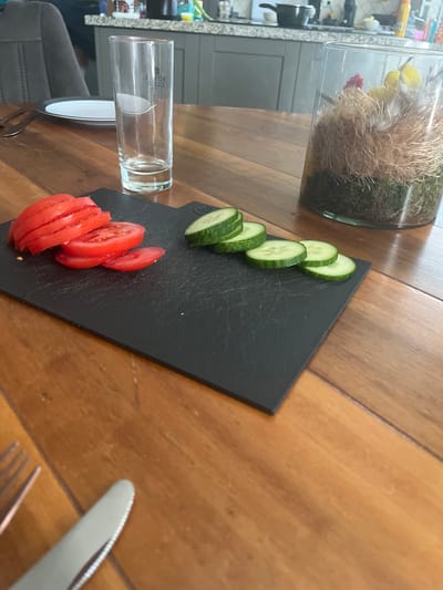 The image depicts a close-up, eye-level view of a wooden table with sliced tomatoes and cucumbers on a black cutting board. A clear glass, a plate with a black rim, and cutlery are also visible on the table. In the background, a kitchen counter with various items and a decorative glass container with what appears to be moss and decorative items are present. The lighting suggests an indoor setting. The location is specified as Uithuizen, Netherlands.