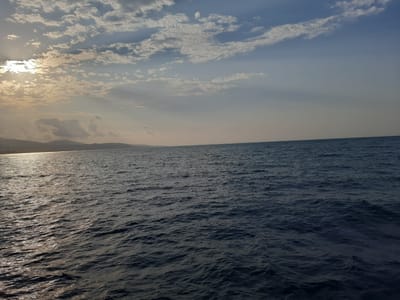 The image depicts a wide expanse of the sea under a cloudy sky. The sun is partially visible through the clouds on the left side of the frame, casting a bright reflection on the water. Gentle waves are visible on the surface of the sea. In the distance, a faint outline of land with hills can be seen. The sky is a mixture of blue and grey clouds, with rays of sunlight piercing through in some areas. This scene is likely captured from a vantage point within or near the city of Atakum, Turkey, given the context provided. No people or specific objects are clearly discernible.