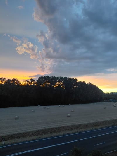 The image depicts a rural landscape scene at dusk, with a field of hay bales in the foreground. A dark treeline runs across the mid-ground, silhouetted against a vibrant sunset sky. The sky is filled with dramatic clouds, ranging from bright white and orange near the horizon to dark grey and blue overhead. A paved road with white markings runs along the bottom of the image. The hay bales are round and scattered across the harvested field. The overall impression is one of a serene, natural setting during twilight hours. There are no people or animals visible in the image. The location is identified as Ecublens, Switzerland.