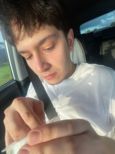 A young male with dark, messy hair and light skin is sitting in the passenger seat of a car, looking down at a small white object in his hands. He is wearing a white t-shirt with an Adidas logo. The sun is shining, casting shadows on his face and shirt. The background shows a glimpse of a suburban or rural environment through the car window, with houses and trees visible under a bright blue sky with scattered clouds. The interior of the car is dark, with a visible headrest and part of a seatbelt. The image appears to be taken outdoors, during the daytime, possibly in the city of Gavrilovka, Russia, based on the provided context.