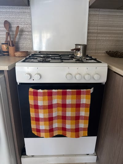 A white gas stove with a black oven door is shown in a kitchen. A red, yellow, and white checkered towel hangs from the oven door handle. On top of the stove, there is a stainless steel pot with a dark brown handle, and several wooden kitchen utensils are in a wooden holder on the counter to the left. The kitchen counter is a light beige color, and the backsplash is made of gray and beige tiled material. To the right of the stove, a decorative woven bowl is visible on the counter. The overall scene is a typical kitchen setting.