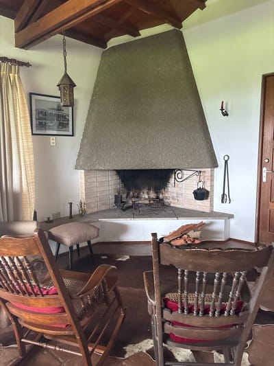 The image shows an interior scene of a room, possibly a living room, featuring a large fireplace with a textured gray exterior and a brick interior. Above the fireplace are fireplace tools, including tongs and a cooking pot hanging on a wrought iron stand. To the right of the fireplace, there are pieces of wood stacked next to the mantel. Two wooden rocking chairs with decorative spindles and red cushions are situated near the fireplace. A smaller upholstered chair is also visible. A black-and-white framed picture hangs on the wall next to the fireplace, below a hanging lantern. Drapes are partially visible. The location in Aucallama, Peru, is not evident from the image's content.
