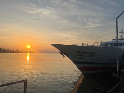 A boat is docked at the pier at sunset. The sun is low on the horizon, reflecting in the water and creating a warm glow. The boat's bow is visible, with Thai script and "SANG LAEM" painted on it. Palm trees on land are visible in the distance. The sky is filled with clouds.