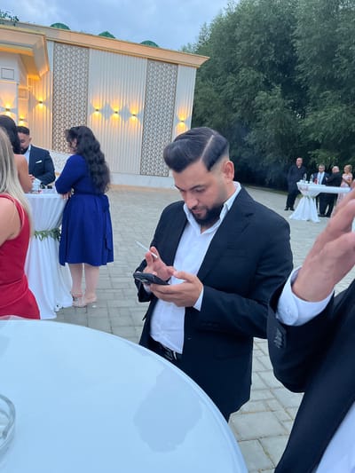 In Satu Nou, Romania, a man in a black suit and white shirt is captured in a candid outdoor setting, possibly at an event or reception. He is looking down at his mobile phone, which he holds in his right hand. A lit cigarette is held between his fingers. His left hand is also near the phone. The man has a neatly styled dark haircut and a beard. In the foreground, a portion of a white round table is visible. Behind him, other individuals and elements of the event space are present, including a white tent-like structure with decorative lights and a backdrop of trees. The ground appears to be paved with interlocking bricks.