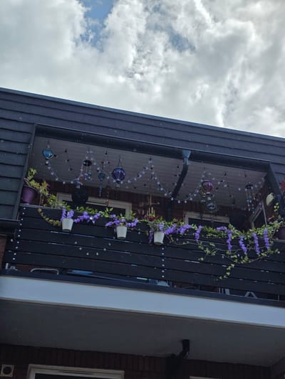 The image displays a decorated outdoor balcony or patio area, viewed from a low angle looking upwards, and is presented in a portrait orientation (rotated 90 degrees clockwise from its natural viewing angle). The foreground features a dark wooden railing or fence that runs diagonally across the frame.
Along the railing, artificial purple flowers and green vines are draped, adding a decorative touch. Several white flower pots are attached to the railing, seemingly holding some of these purple blooms, and a larger purple pot with green foliage is also visible. Behind the railing, reflecting in what appears to be a large window or glass door, is a display of string lights featuring star shapes and spherical, translucent ornaments, possibly hot air balloon or lantern designs, adding a whimsical element. A dark, possibly black, rain gutter or pipe is visible on the building structure to the left of the balcony.
To the right of the balcony structure, through the glass and beyond, a brick wall of a building is visible, along with a window framed in white. The background, above and to the left of the balcony, shows a cloudy sky with patches of brighter blue, indicating daytime. There are no people visible, and no specific activity is taking place other than the presentation of a decorated outdoor space. No visible text is present in the image. The scene suggests a personalized and welcoming exterior environment.