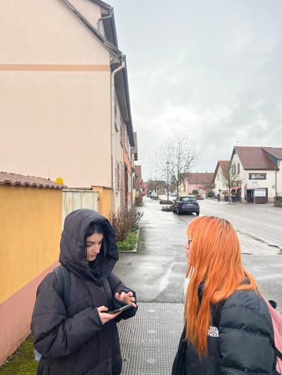 The image depicts an outdoor scene on a wet, overcast day in Meistratzheim, France. In the foreground, two young females are present on a concrete sidewalk. The female on the left wears a black hooded puffer jacket, has dark hair, and is looking down at a black smartphone held in both hands, actively interacting with its screen. A black backpack strap is visible on her left shoulder. To her right, the second female has bright orange hair with dark roots, wears glasses, and a black puffer jacket with a partially visible "THE NORTH FACE" logo. She is turned to her left, looking towards the first female. A pink backpack strap is visible on her right shoulder.
The immediate environment includes a section of a metal grate on the sidewalk between the two individuals. To the left, a low yellow-orange wall with a terracotta-tiled coping borders a light beige building with a single horizontal peach stripe. The street beyond the sidewalk appears wet, indicating recent precipitation. A dark purple Renault Clio is parked on the street in the mid-ground. Buildings line both sides of the street, featuring various colors (beige, orange, white) and red-tiled roofs. A bare deciduous tree stands on the sidewalk further down the street. On the right side of the street, a white building displays signage, including "CRÊPES GLACES" and "DISTRIBUTEUR." The sky is uniformly grey and overcast.