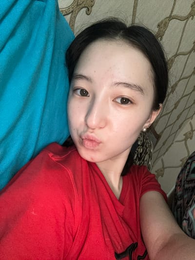 A young East Asian woman with dark hair and light skin is captured in a close-up, selfie-style photograph. She is wearing a red t-shirt and a pearl earring in her visible right ear. Her lips are pursed in a kissing gesture. The background consists of a textured wallpaper with a nature-inspired pattern and a large piece of bright blue fabric. The lighting appears to be natural and soft. The image is taken indoors, possibly in a bedroom, given the presence of the fabric and wallpaper. The exact location in Nookat, Kyrgyzstan cannot be determined from the visual information provided.
