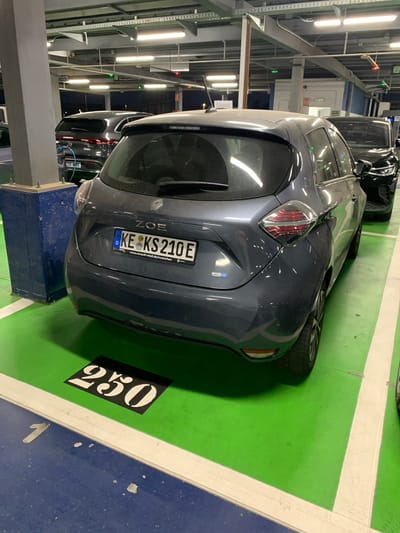 The image shows the rear of a grey Renault Zoe electric car parked in a brightly lit multi-story car park. The car has a German license plate that reads "KE KS 210E". The parking spot is marked with green flooring and a white line, and a black square with the number "250" is painted on the ground in front of the car. In the background, other cars and parking infrastructure, including lights and a pillar, are visible. The overall environment suggests an indoor parking facility in a city.