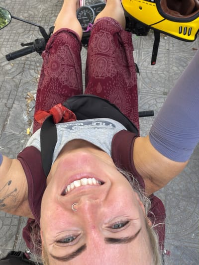 In this overhead, full-body selfie taken from the perspective of someone sitting on a motorcycle, a woman with blonde hair and a nose ring smiles directly at the camera. She is wearing a burgundy patterned skirt, a gray t-shirt, and a burgundy tank top. A red bandana is tied around her neck. A yellow helmet with brown trim sits on the back of the motorcycle. The motorcycle is parked on a tiled surface, likely a street in Đồng Hới Ward, Vietnam, as indicated by the context. The background shows a portion of the motorcycle's dashboard and a mirror.