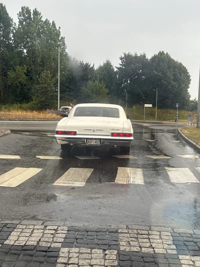 The image depicts a white Chevrolet Impala from the mid-1960s, with a Swedish license plate reading "AGP 482", parked on a wet road at a pedestrian crossing. The car is facing away from the camera, with its taillights illuminated. It is raining, as evidenced by the wet road surface, reflections, and visible raindrops. In the background, there are trees, streetlights, and a sign that reads "Karlsgatan". The road is paved with cobblestones in the foreground and asphalt further back. The sky is overcast, suggesting a cloudy or rainy day. There are no people clearly visible interacting with the car, though a person's hand is raised in another car in the distance.