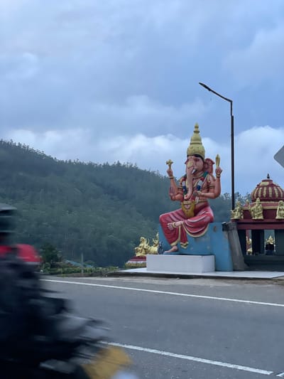 The image depicts a roadside statue of the Hindu god Ganesha in Seetha Eliya, Sri Lanka. The statue is positioned on a concrete base next to a road with white lines. A green, forested hillside rises in the background under a cloudy sky. A streetlight is visible to the left of the statue. A portion of a building with a tiered, gold-colored roof is visible above the statue. The image appears to be taken from a moving vehicle, as evidenced by the blurred foreground.