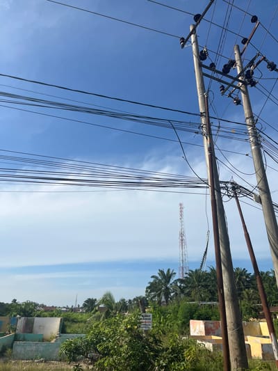 This is a ground-level, outdoor, daytime photograph taken in Tanahterban, Indonesia. The image captures a view of utility poles with numerous overhead wires against a clear blue sky with some wispy white clouds. In the background, a tall, red and white communication tower is visible, surrounded by lush green vegetation, including palm trees and smaller trees. Some partially constructed or dilapidated concrete structures are also present in the mid-ground, with signs of paint in various colors. The overall scene depicts a rural or semi-urban environment with infrastructure and natural elements.