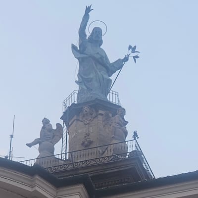 The image shows a statue of Jesus Christ on top of a building in Calcinate, Italy. The statue is made of stone and depicts Jesus with his right hand raised in a blessing gesture and holding a branch in his left hand. He is wearing a halo around his head. Below the statue of Jesus, there are several smaller statues of angels. The building appears to be made of stone and has a decorative railing. The sky is overcast and hazy, suggesting it might be a foggy or cloudy day. The overall scene is solemn and religious in nature, with the statue of Jesus serving as the focal point.