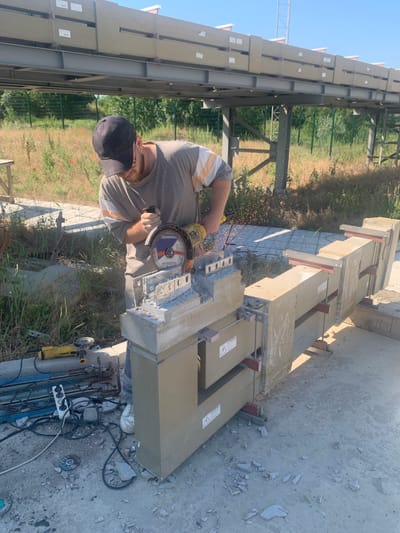 A man wearing a black baseball cap, a grey t-shirt with striped sleeves, and white sneakers is using a yellow and black angle grinder to cut through a concrete structure. Sparks are flying from the grinding process. The concrete structure appears to be part of a larger installation, with multiple beige-colored rectangular blocks lined up on a metal frame. The scene is outdoors, with dry grass and vegetation in the background. There is a fence and some trees behind the grass. Other tools and materials, including extension cords, a power strip, and metal rods, are scattered on the ground around the work area. The overall environment suggests a construction or industrial site. The location is identified as Pivdennoukrainsk, Ukraine.