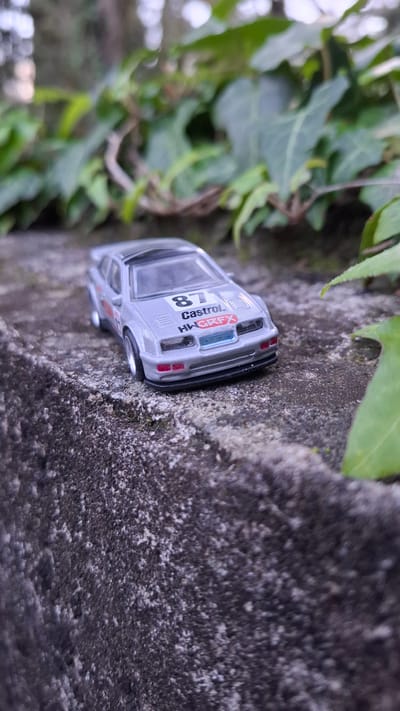 A silver toy car, labeled "8" and "Castrol HWGRFX", sits on a textured, gray concrete surface, seemingly the top of a low wall. Green foliage and ivy are visible to the left of the wall and behind the car. The scene appears to be outdoors. The location of Podbadac, Montenegro is not apparent in the image.