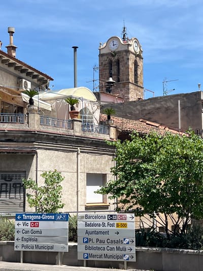 The image shows a street scene in Mollet del Vallès, Spain, under a clear blue sky. In the foreground, two multi-panel street signs are visible. The left sign directs to "Tarragona-Girona AP-7," "Vic C-17," "Can Gomà," "Església," and "Policia Municipal." The right sign points towards "Barcelona C-33 C-17," "Badalona-St. Fost B-500," "Ajuntament," "Pl. Pau Casals," "Biblioteca Can Mulà," and "Mercat Municipal." Behind the signs, a building with balconies and potted plants is visible, and further in the background, a stone clock tower with bells rises against the sky. Lush green trees and foliage are present on the right side of the frame.