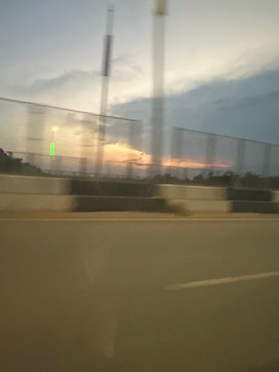 The image depicts a blurred view of a road in Kunkwaba, Nigeria, likely taken from a moving vehicle. The foreground shows a section of asphalt road. Beyond the road, there is a concrete barrier with alternating black and white sections, likely a roadside divider. Behind the barrier, a fence runs horizontally across the image. The sky above is cloudy with hints of sunset colors, suggesting it is either dawn or dusk. A tall, slender structure, possibly a light pole or communication tower, is visible in the sky, blurred by motion. A bright yellow light is also visible near the fence, possibly from a streetlamp. A green rectangular object is also visible on the fence. The overall impression is of a transitional landscape, possibly on the outskirts of a city, characterized by infrastructure and the natural elements of the sky.