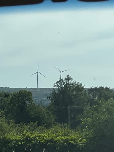 The image shows a distant view of two wind turbines silhouetted against a bright, overcast sky. They are positioned on a hill with a dark horizon line. In the foreground, there are dense green trees and bushes. A telephone pole with wires is visible amongst the trees. The scene appears to be a rural landscape. The city of Derrylin, United Kingdom, is not depicted in this image, as it is a small village in County Fermanagh, Northern Ireland.