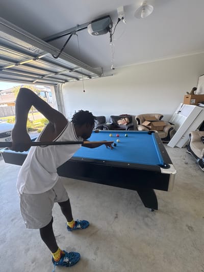 A young person, appearing to be a male with dark skin and dreadlocks or braids, is playing pool inside what appears to be a garage in Officer South, Australia. He is positioned at a blue-felt pool table, holding a cue and aiming at several pool balls, including a white cue ball, a yellow ball, a red ball, and a black ball. He is wearing a white sleeveless top with black trim, grey shorts, blue athletic shoes, and yellow socks.
The setting is a well-lit garage, with a concrete floor and white walls/ceiling. An automatic garage door opener motor is mounted on the wall/ceiling, with wires connected to it. A segmented garage door is partially visible, indicating an opening that lets in natural daylight, suggesting it is daytime. Outside the garage, some greenery and what looks like a residential street can be glimpsed, further confirming the domestic setting. In the background, stored items such as cardboard boxes and what appear to be household furniture items are visible, consistent with a garage space. The overall impression is one of casual recreation in a home environment. No discernible text is visible on any objects within the frame.