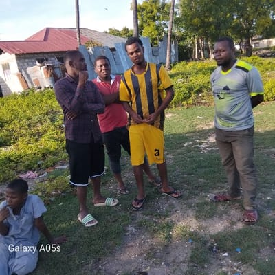 The scene is located in Nungwi, Tanzania, at coordinates -5.726 latitude and 39.301 longitude. Four men are standing on a grassy area in front of a building under a cloudy sky. The man in the middle, wearing a black and yellow jersey, is looking down at his hands. The man to his left is wearing a red shirt and black shorts. He is looking to the right. The man on the far right is wearing a gray and black shirt. He is looking at the camera. The man to his left is wearing a plaid shirt and black shorts. He is looking at the camera. The man on the far left is sitting down and is looking at the camera. He is wearing a gray shirt and black pants. There is a partially visible building in the background and vegetation in the foreground. The ground is covered in grass and dirt.