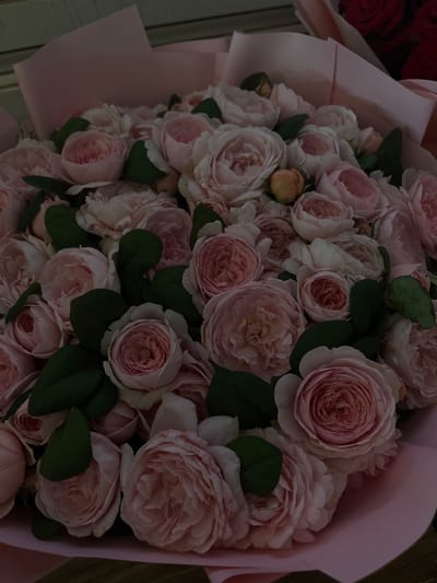 This is a close-up, overhead shot of a large bouquet of light pink roses. The roses are densely packed and vary in bloom, with some appearing as tight buds and others fully open with ruffled petals. A few small, peach-colored buds are also visible amongst the pink blooms. Dark green leaves are interspersed throughout the bouquet, providing a contrast to the soft pink of the flowers. The bouquet is partially wrapped in light pink paper, which is visible in the foreground and on the right side of the image. The background is out of focus, but appears to be a wooden surface. The lighting is soft and diffused, suggesting an indoor setting or an overcast day. The overall impression is one of romantic elegance and natural beauty. No text is visible in the image.