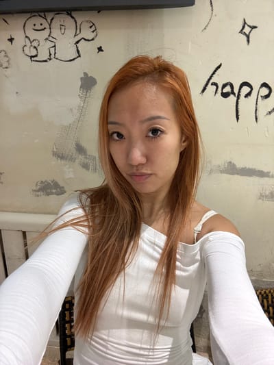 A forensic examination of the image reveals a medium shot of an East Asian female, approximately 20-30 years old, with long, reddish-brown hair and a light complexion, looking directly at the camera. She has visible makeup, including eyeliner and possibly false eyelashes. Her expression is neutral. She is wearing a white, long-sleeved, off-the-shoulder top or dress, which appears ribbed or ruched in sections. A white bra strap is visible on her right shoulder. The woman is positioned against a cream-colored, textured wall that shows signs of wear, including patches of gray paint or plaster. Black, hand-drawn graffiti adorns the wall, including two cartoon-like figures (one with a smiley face and a thumbs-up, the other with an open mouth and outstretched arm), several small star shapes, and the word "happ" (likely part of "happy"). To the lower left, a portion of a white, slatted object, possibly a bench or railing, is visible. The scene is located in Lishan District, China.