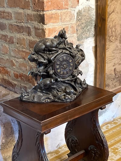 A dark, ornate antique clock featuring sculpted animals sits on a polished wooden table in Trakai, Lithuania. The clock's base depicts several animals, possibly dogs or wolves, in dynamic poses. The clock face itself is circular with Roman numerals. The table has carved, ornate legs and is placed against a brick wall. To the right of the clock, a portion of a framed tapestry or painting is visible. The floor has a yellow and white striped pattern with a floral motif.