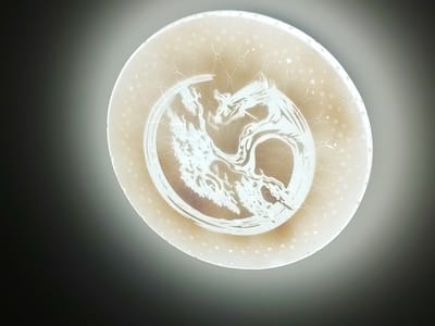 The image shows a close-up, overhead view of a circular, translucent plate with a golden-brown hue. The plate is decorated with a white, etched design that appears to be two intertwined dragons. The dragons are stylized and rendered in a traditional, Asian-inspired art style, with flowing lines and intricate details. Around the dragons, the plate is adorned with a pattern of small, light-colored stars and faint, geometric line work resembling constellations. The plate is illuminated from above, creating a soft glow and casting a subtle shadow on the dark background. The background is a gradient of dark gray to light gray, suggesting the plate is placed on a flat surface. There is no indication of the city of Сухарево, Russia, in the image itself.