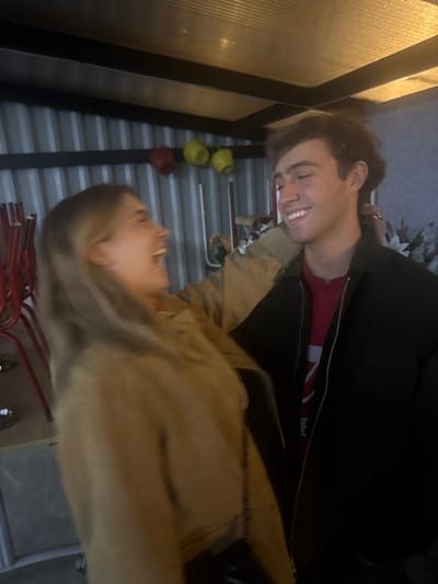 A candid, medium-shot captures two individuals, one male and one female, in an indoor or sheltered outdoor setting within Décines-Charpieu, France. The female, positioned on the left and slightly out of focus, has light-colored hair and wears a light brown or tan jacket. Her head is turned to the right, mouth open in an apparent laugh or exclamation, with her right arm extended towards the male. The male, positioned on the right and in sharp focus, has short, wavy brown hair and is smiling broadly, looking down towards the female. He wears a dark, collared jacket over a red hooded sweatshirt with white graphics. His left arm is visible, appearing to be around the female's back. The background consists of a vertically corrugated, light grey metal wall. A dark horizontal beam crosses the upper portion of this wall, from which one red and two yellow spherical objects, possibly decorative gourds or lights, are suspended. A translucent, possibly corrugated plastic, ceiling with warm overhead lighting is visible at the top. To the far left, a stack of red metal chairs is partially visible, along with a silver bowl on the ground. To the right, white artificial floral decorations and a white cylindrical object (possibly a candle) are present. The scene is illuminated by warm, artificial light, highlighting the subjects' expressions of mirth and interaction. The overall environment suggests a casual gathering space, possibly a converted shed, event tent, or community area.