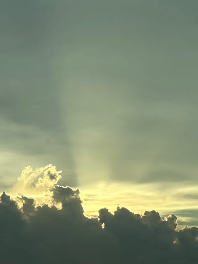 The image displays a dramatic sky scene with a band of bright yellow light near the horizon, partially obscured by dark, heavy clouds. Above this, lighter, wispy clouds catch the sunlight, creating a contrast of light and shadow. A vertical shaft of light, possibly a sunbeam, pierces through the upper, greenish-grey sky. The overall impression is of a stormy or sunset sky. The image is a photograph taken from an elevated perspective, looking upwards. There are no people, structures, or recognizable landmarks visible that would indicate a specific location within Fort Myers, United States of America. The focus is entirely on the atmospheric conditions.