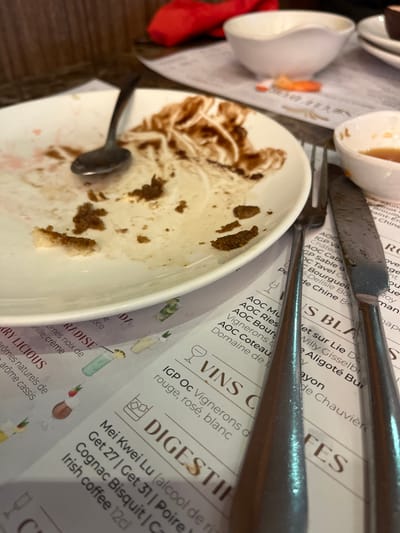 The image shows a cluttered dining scene. A mostly empty white plate with remnants of a chocolate dessert sits alongside silverware. Next to the plate is a menu featuring wine and digestif options. The menu lists a selection of wines and digestifs, like "Mei Kwei Lu", "Cognac Bisquit", "Irish Coffee", and various AOC wines. There is also an empty white bowl and glass on the left. The photo suggests the end of a meal with the focus on the leftovers and menu.