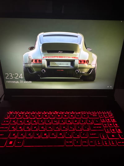 The image shows a laptop with a screensaver of a vintage green Porsche 911. The time displayed on the screen is 23:24, and the date is Friday, July 31st. The laptop keyboard is illuminated with red backlighting. The surrounding environment is dark, suggesting it is nighttime or the room is dimly lit. The image appears to be taken in a personal setting, possibly a home or office, and does not provide any contextual clues to suggest it is located in Nizhniy Kuranakh, Russia.
