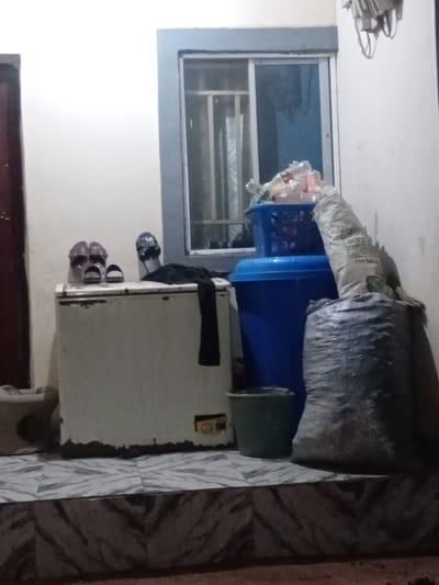 The image depicts an indoor scene, likely a storage area or a less organized part of a dwelling, in Pantinapu, Nigeria. The primary subjects are various objects. In the background, a window with a blue-tinted pane is visible. In front of the window, a large blue plastic bin is filled with what appear to be plastic bottles and other miscellaneous items. Resting on top of the blue bin is a large, grey, tarp-like bag, possibly containing more stored goods. To the right of the blue bin, a small, dark green rectangular object is placed.

In the foreground, a light-colored, rectangular appliance, possibly a small refrigerator or freezer, is positioned. A black piece of fabric is draped over the top of this appliance, and below it, a pair of dark-colored sandals and another pair of brown sandals are placed on what appears to be a shelf or the floor. The floor in the right portion of the image is tiled with a grey and white pattern. The overall impression is one of clutter and everyday utility, rather than a curated display. There is no clear indication of the time of day or weather, and the lighting suggests an indoor environment. No people are visible in the image. No text is clearly legible.