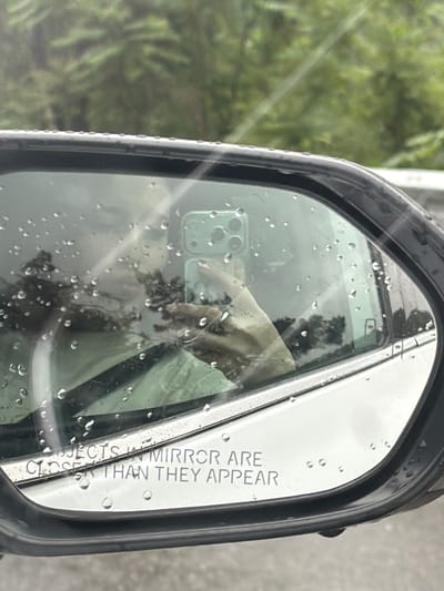 The image shows a driver's side mirror of a vehicle on a rainy day, reflecting a mobile phone being used to capture the scene. Water droplets are visible on the mirror's surface. The mirror's inscription reads "OBJECTS IN MIRROR ARE CLOSER THAN THEY APPEAR." The background suggests a wooded area, likely visible from the vehicle's location. The location of the scene in Grigalati, Georgia is not possible to verify from the image.