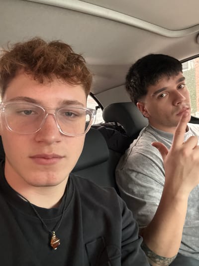 Two young men are depicted in a selfie taken inside a vehicle, likely a car or bus, in Ramos Mejía, Argentina. The image is oriented sideways, suggesting it was taken with the camera held horizontally.
In the foreground, a young man with light skin, reddish-brown curly hair, and clear-framed glasses looks directly at the camera with a neutral expression. He is wearing a dark top and a necklace with an amber-colored, possibly hexagonal or teardrop-shaped pendant, which hangs near his chest.
Behind him, positioned slightly higher and to the left, is another young man with dark hair, wearing a light grey t-shirt. He has a band-like tattoo visible on his upper left arm. This man looks towards the camera with a slightly playful or curious expression, making a gesture with his right hand where his index finger is either touching his mouth or pointing upwards.
The interior of the vehicle features light grey panels and dark grey headrests and seats. Through a window in the background, a glimpse of an outdoor scene is visible, including what appears to be a building with a reddish roof and some greenery, indicative of a daytime setting, possibly in a suburban or residential area. The lighting is soft and diffused, suggesting an overcast day or indirect sunlight. The overall atmosphere is casual. No visible text is present in the image.