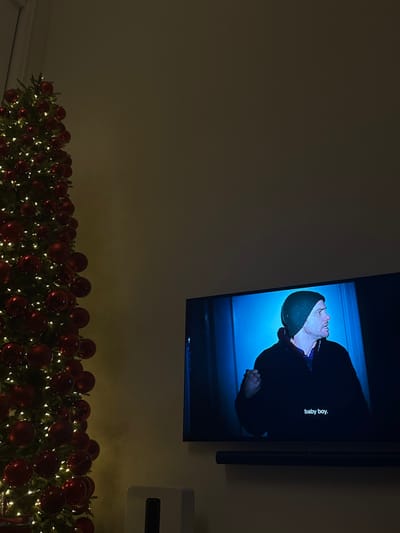 A television displays a blue-toned image of a man wearing a dark beanie and jacket, the subtitles reading "baby boy." Beneath the TV, a festive Christmas garland with red ornaments and small white lights decorates the scene. The wall is a muted beige color. An off-white electronic device is mounted to the right of the television. The lighting in the room is dim, creating a cozy and slightly mysterious ambiance.