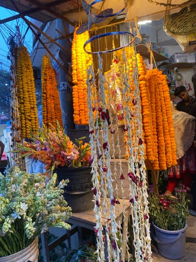 This image captures a detail-rich flower stall scene in Narayanganj, Bangladesh. The foreground and mid-ground are dominated by an extensive display of fresh flowers. Vertically suspended are numerous long garlands of vibrant orange and yellow marigolds, alongside intricate strands of white tuberose flowers interspersed with dried reddish-pink petals, hanging from blue circular frames.

On the left, a white bucket contains a large arrangement of white tuberose buds. Adjacent to this, a black bucket holds an assortment of colorful gladiolus flowers in various shades of orange, pink, and yellow. Towards the lower right, a grey bucket presents purple and pink chrysanthemum-like flowers with green stems. The stall features visible wooden planks and posts, forming a makeshift structure.

In the mid-ground right, a young man with dark hair, wearing a dark short-sleeved shirt, is visible from the chest up, looking downwards. To his lower right, the lower legs and feet of another individual are visible, clad in red trousers and a patterned garment, possibly a saree. In the far left background, a person in a white shirt is partially obscured by the flower displays.

The background on the left reveals an outdoor setting with crisscrossing overhead wires against a light blue sky. The background on the right shows an indoor or covered area with shelving, various boxes (one labeled "Oasis"), a security camera mounted on the wall, and signs with Bengali script. The scene is illuminated by both natural ambient light from the left and artificial lighting fixtures within the stall structure.