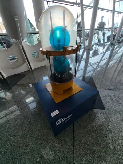 A tilted medium shot captures a display piece, likely located inside an airport terminal, given the reflective glass ceiling and exterior views visible through the windows. The display features a vintage-style aviation beacon encased in a clear dome, containing two blue bulbs. The beacon is mounted on a yellow base, which is in turn attached to a dark blue pedestal.

The pedestal has text on its side, which appears to reference "Aeroporto" (airport in Portuguese) and "Porto," indicating a connection to Porto Airport. The location of the airport is in Moreira, Portugal. The flooring consists of polished speckled granite tiles. The display is adjacent to a section of wall with porthole-like windows. There are no people visible in the shot.