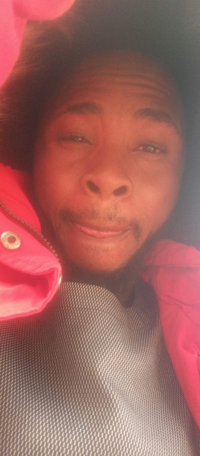 The image is a close-up, high-angle selfie of a person with dark skin. The person is looking directly into the camera with their head slightly tilted. Their eyes are open, with a slight squint, and their mouth is slightly open, revealing a bit of their tongue. There is some facial hair visible above their upper lip and on their chin. The overall facial expression appears somewhat playful or quirky.
The person is wearing a bright, possibly fuchsia or vibrant red, hooded jacket or sweatshirt. A silver eyelet or snap fastener is clearly visible on the left side of the jacket, near the shoulder area. Underneath the jacket, a grey or dark-colored shirt with a distinctive, finely patterned texture is visible. The pattern consists of small, repeating rectangular or dash-like shapes arranged in a grid or woven pattern, creating a textured surface. The hood of the jacket seems to be up, framing the person's head and contributing to the dark areas surrounding their face.
The lighting in the image is somewhat warm and soft, casting a reddish glow across the person's face, possibly due to ambient light or a filter. The image has a noticeable grain or noise, particularly evident in the darker areas and skin tones. The background is indistinct and appears dark, blending with the interior of the hood, making it impossible to discern any specific setting. No text is visible in the image.